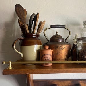 May include: A rustic kitchen shelf displays a collection of vintage items. A brown and cream ceramic jar holds wooden spoons. A copper kettle, a small orange spice jar labeled "CURRY", and glass jars complete the arrangement.