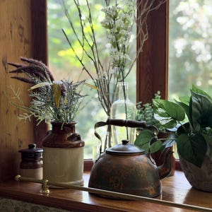 May include: A copper-toned tea kettle with a dark patina sits alongside a cream and brown ceramic milk jug filled with dried grasses, a clear glass bottle with white floral stems, and a green leafy plant on a wooden windowsill.