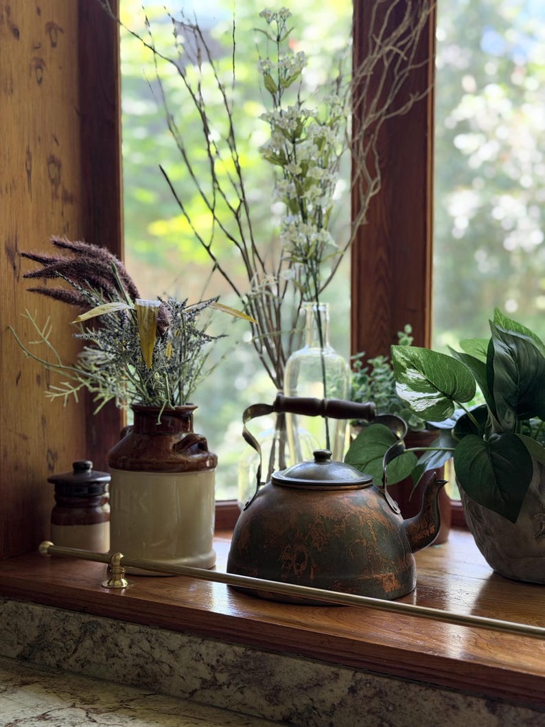 May include: A copper-toned tea kettle with a dark patina sits alongside a cream and brown ceramic milk jug filled with dried grasses, a clear glass bottle with white floral stems, and a green leafy plant on a wooden windowsill.