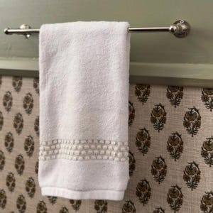 May include: A white cotton hand towel with a textured pom-pom trim detail hanging on a polished silver-toned metal towel rail. The towel features a soft, absorbent fabric construction with a decorative band of small, raised circular accents.