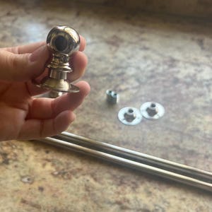 May include: A polished silver curtain rod finial with a spherical top and ornate base. The image also shows two silver curtain rods, mounting hardware, and a hand holding the finial.