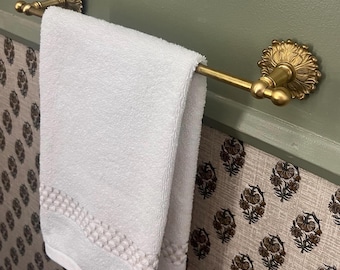 May include: A white cotton hand towel with a textured woven border hangs over a horizontal brass towel rail. The rail features ornate, circular wall-mounted brackets with detailed floral-inspired engravings on each end, finished in a matte gold tone.