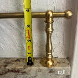 May include: A brass towel rail featuring a cylindrical rod and a decorative pedestal base. A yellow measuring tape is positioned vertically against the pedestal, showing a height of approximately 10 cm from the base to the top of the rod.