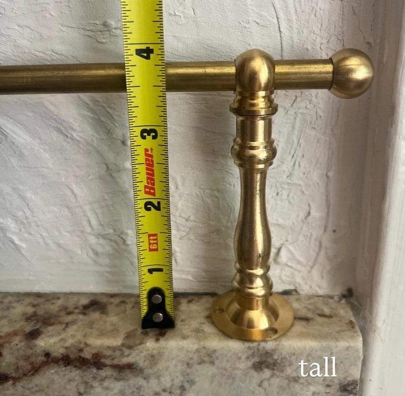 May include: A brass towel rail featuring a cylindrical rod and a decorative pedestal base. A yellow measuring tape is positioned vertically against the pedestal, showing a height of approximately 10 cm from the base to the top of the rod.