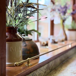 May include: A two-toned brown and cream ceramic milk jug with embossed text, holding a bouquet of dried grasses and foliage. A polished brass rail is mounted horizontally along the edge of a wooden window sill next to a dark metal kettle.