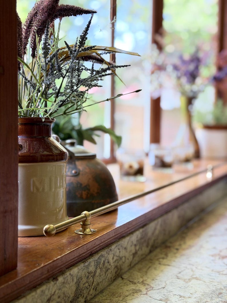 May include: A two-toned brown and cream ceramic milk jug with embossed text, holding a bouquet of dried grasses and foliage. A polished brass rail is mounted horizontally along the edge of a wooden window sill next to a dark metal kettle.