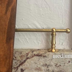 May include: A brass rail with a cylindrical rod and a decorative pedestal base featuring a rounded finial end. The metal hardware is mounted horizontally and includes the word medium printed in white text on the surface below the rod.