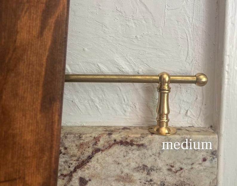 May include: A brass rail with a cylindrical rod and a decorative pedestal base featuring a rounded finial end. The metal hardware is mounted horizontally and includes the word medium printed in white text on the surface below the rod.