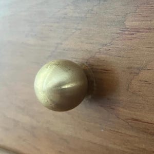 May include: A round, brass-colored cabinet knob. The knob is a solid sphere with a smooth, polished surface. The knob is mounted on a wooden surface with a natural wood grain.