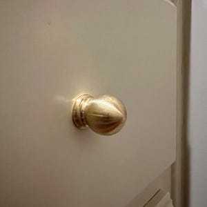 May include: A polished brass cabinet knob with a rounded, acorn-like shape and a tiered circular base, mounted onto a flat, off-white drawer front. The metal surface reflects light, highlighting its smooth, curved, and symmetrical design.
