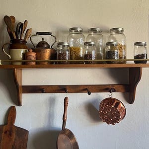 May include: A wooden wall shelf with a brass rail, featuring a collection of glass jars filled with dry goods, a ceramic pitcher with wooden utensils, a copper kettle, and a copper mould hanging from one of the four hooks on the lower rack.