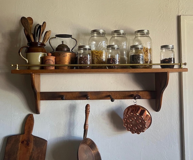 May include: A wooden wall shelf with a brass rail, featuring a collection of glass jars filled with dry goods, a ceramic pitcher with wooden utensils, a copper kettle, and a copper mould hanging from one of the four hooks on the lower rack.