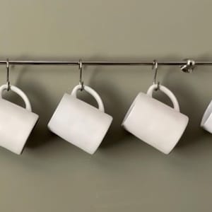 May include: Four plain white ceramic mugs hanging from a horizontal silver metal rail. Each mug is suspended by a silver S-shaped metal hook attached to the rail. The mugs are evenly spaced and feature a simple cylindrical shape with a curved handle.