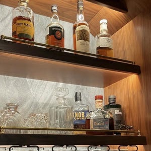 May include: Two wooden shelves with brass railings holding various glass liquor bottles, including Frey Ranch, Springbank, Compass Box, Highland Park, Knob Creek, and George Dickel, alongside a crystal decanter and hanging wine glasses.