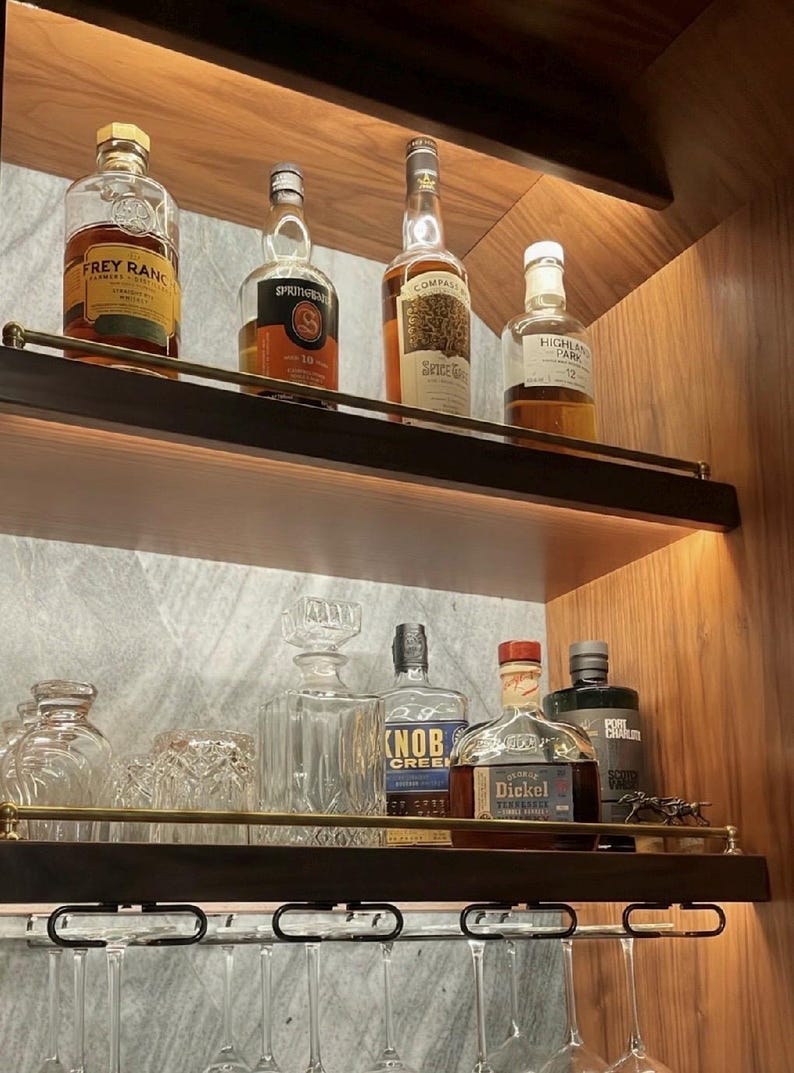 May include: Two wooden shelves with brass railings holding various glass liquor bottles, including Frey Ranch, Springbank, Compass Box, Highland Park, Knob Creek, and George Dickel, alongside a crystal decanter and hanging wine glasses.