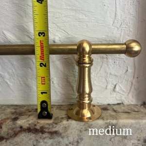 May include: A brass towel bar with a cylindrical rod and a decorative pedestal base, positioned next to a yellow measuring tape showing a height of approximately 9 cm from the base to the top of the rod, with the word medium printed below.