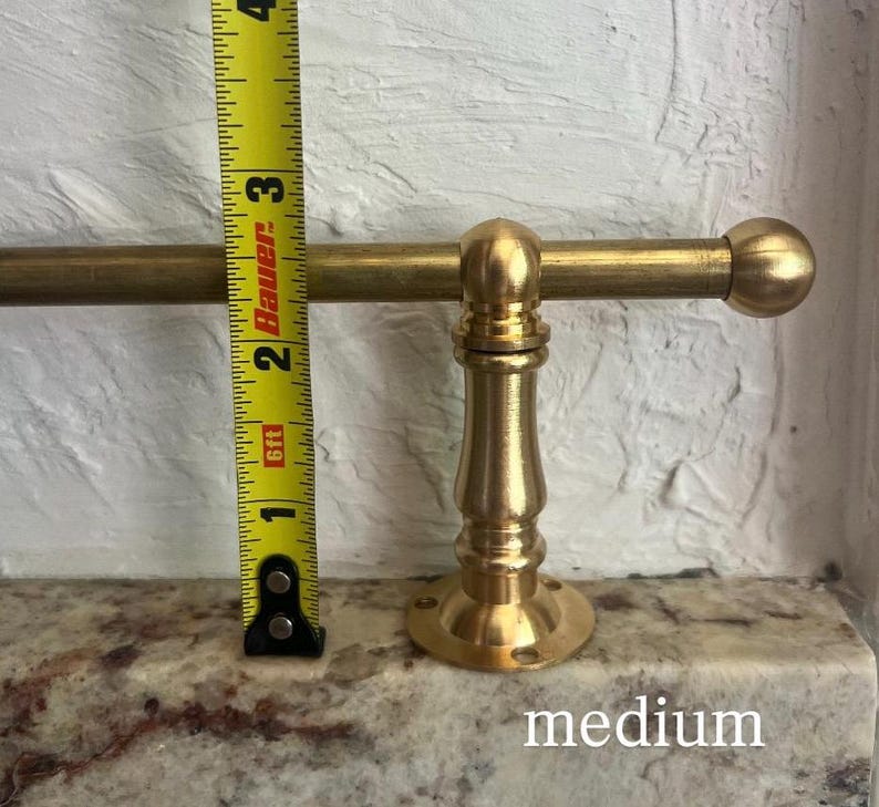 May include: A brass towel bar with a cylindrical rod and a decorative pedestal base, positioned next to a yellow measuring tape showing a height of approximately 9 cm from the base to the top of the rod, with the word medium printed below.