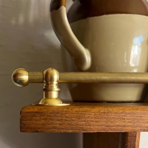May include: A horizontal brass rail with a rounded end cap and a circular mounting bracket attached to the edge of a wooden shelf. The metal has a smooth, matte finish, and the rail is positioned in front of a two-toned brown ceramic pitcher.