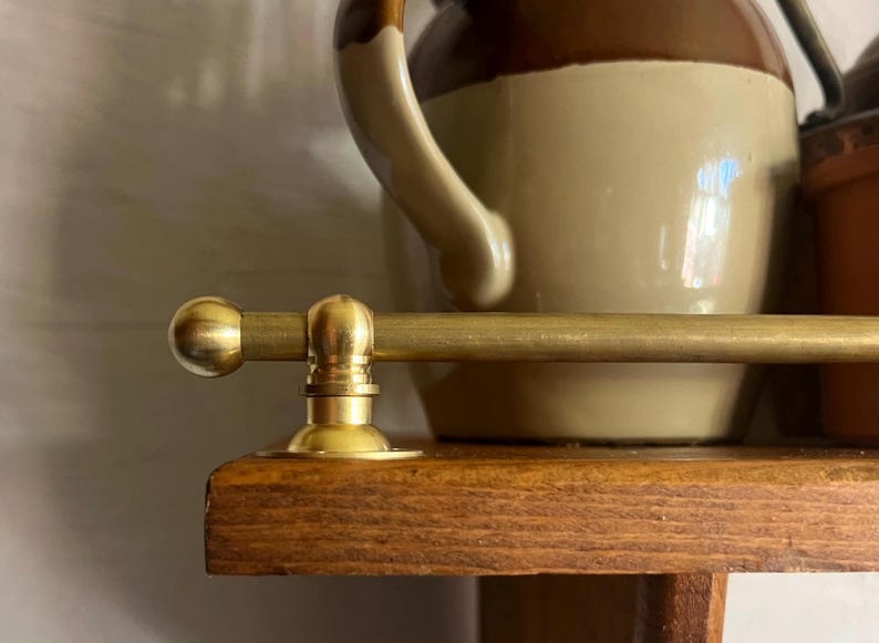May include: A horizontal brass rail with a rounded end cap and a circular mounting bracket attached to the edge of a wooden shelf. The metal has a smooth, matte finish, and the rail is positioned in front of a two-toned brown ceramic pitcher.