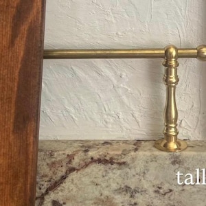 May include: A brass rail with a decorative vertical support post featuring a rounded finial end. The metal hardware is positioned horizontally above a speckled granite surface, with a dark wood panel visible on the left side. The word tall is printed.