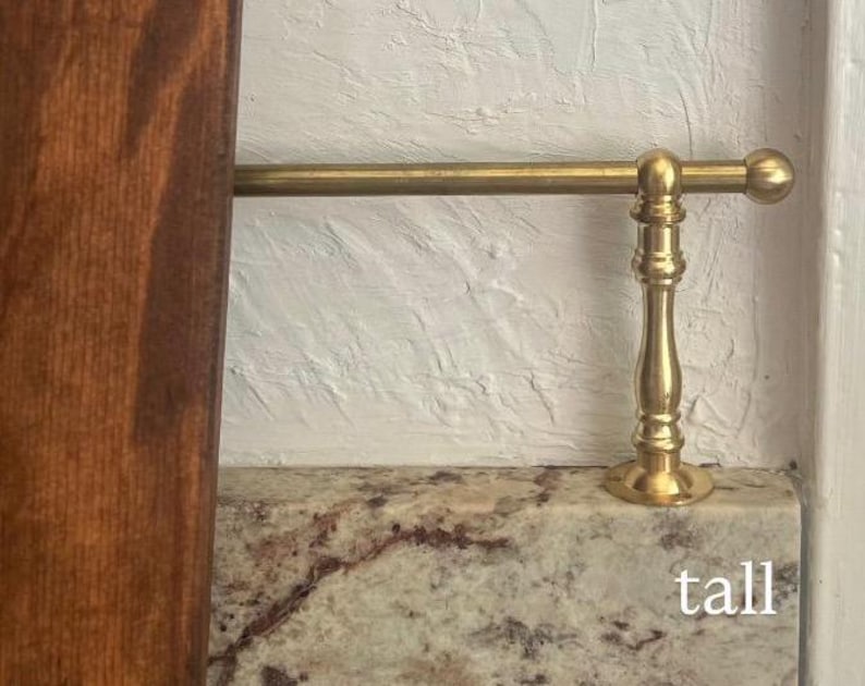 May include: A brass rail with a decorative vertical support post featuring a rounded finial end. The metal hardware is positioned horizontally above a speckled granite surface, with a dark wood panel visible on the left side. The word tall is printed.