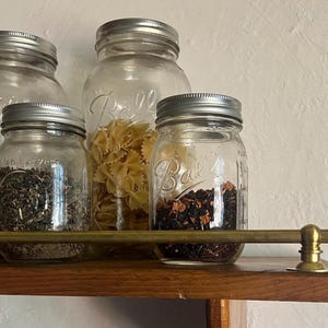 May include: Clear glass Ball mason jars with silver metal lids containing various dry goods like pasta, tea leaves, and herbs, arranged on a wooden shelf behind a horizontal brass rail with spherical end caps and circular mounting hardware.