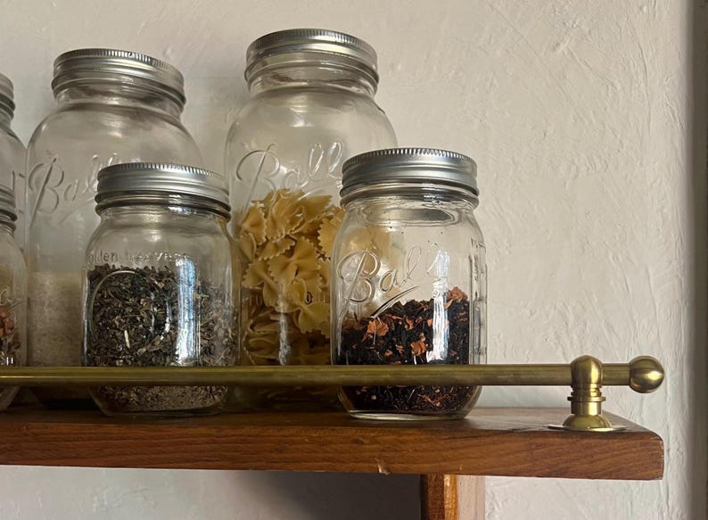 May include: Clear glass Ball mason jars with silver metal lids containing various dry goods like pasta, tea leaves, and herbs, arranged on a wooden shelf behind a horizontal brass rail with spherical end caps and circular mounting hardware.