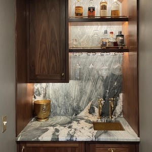 May include: A built-in wet bar featuring dark wood cabinetry, a green and white marble countertop and backsplash, and a small gold sink. Two shelves hold various glass liquor bottles, while stemware hangs from a rack above the sink.