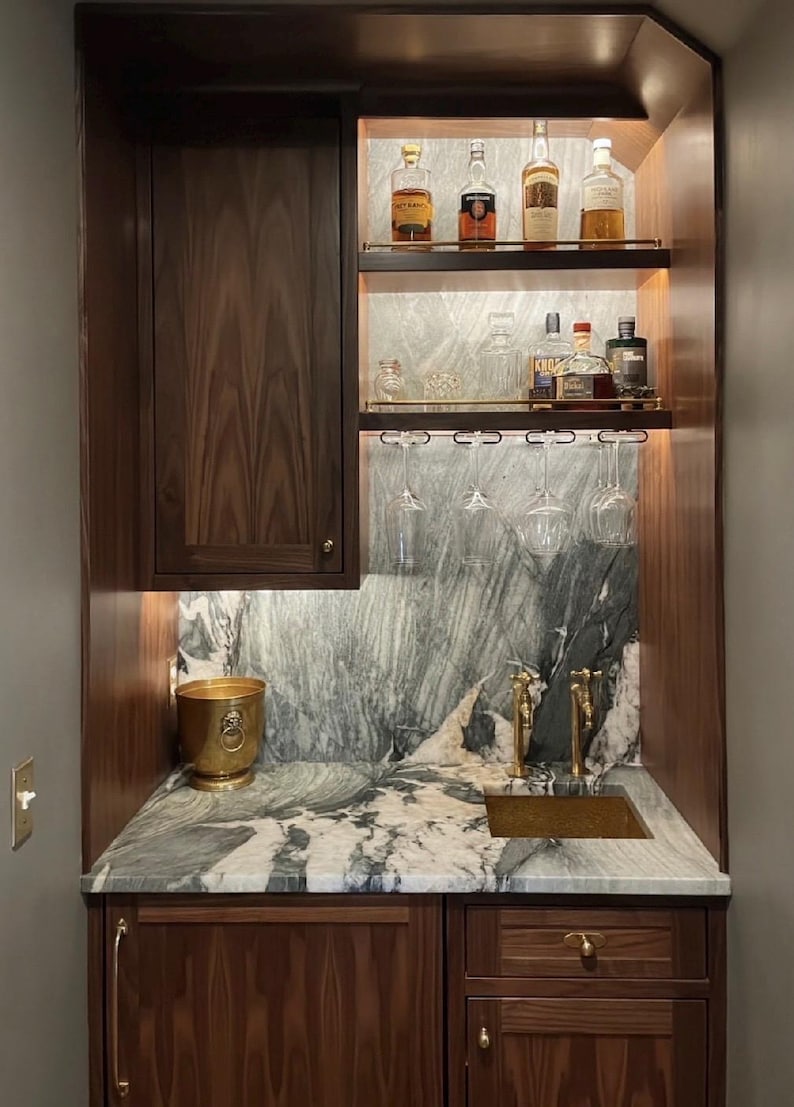May include: A built-in wet bar featuring dark wood cabinetry, a green and white marble countertop and backsplash, and a small gold sink. Two shelves hold various glass liquor bottles, while stemware hangs from a rack above the sink.