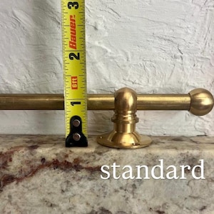 May include: A brass curtain rod with a spherical finial and a circular mounting bracket, positioned next to a yellow measuring tape showing a height of approximately 3.8 cm for the bracket. The word standard is printed below.