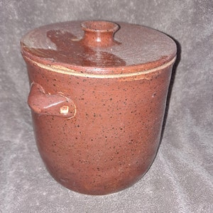 Puede incluir: Un tarro de cer&aacute;mica marr&oacute;n rojizo moteado con tapa y asa. El tarro tiene un cuerpo redondeado y una tapa con un peque&ntilde;o pomo. El asa est&aacute; sujeta al lateral del tarro. El tarro parece hecho a mano.