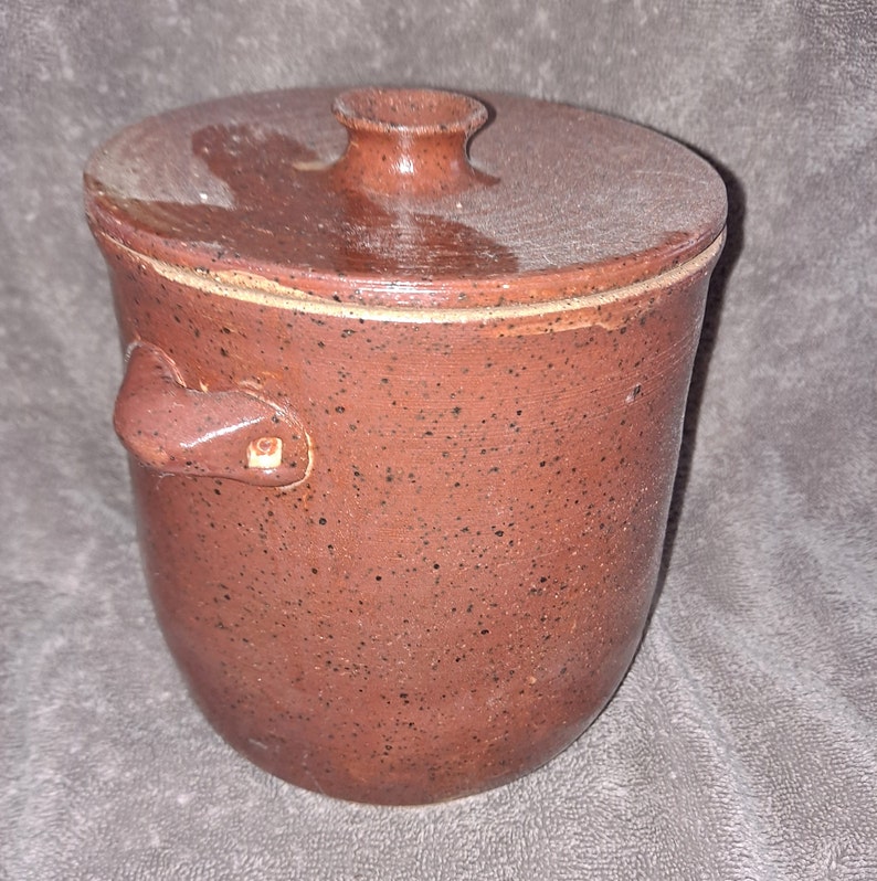 Puede incluir: Un tarro de cer&aacute;mica marr&oacute;n rojizo moteado con tapa y asa. El tarro tiene un cuerpo redondeado y una tapa con un peque&ntilde;o pomo. El asa est&aacute; sujeta al lateral del tarro. El tarro parece hecho a mano.