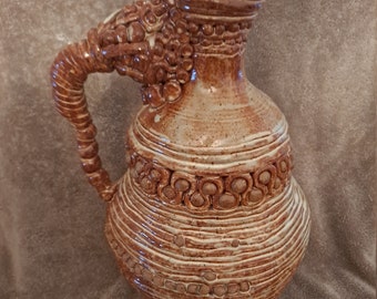 May include: A brown and white ceramic pitcher with a unique, textured design. The pitcher has a wide, round body and a tall, slender spout. The handle is twisted and has a decorative design.