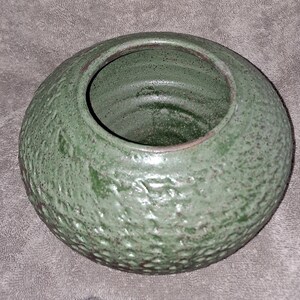 Puede incluir: Un jarr&oacute;n redondo de cer&aacute;mica verde con un exterior texturizado. El jarr&oacute;n tiene una abertura ancha y una superficie ligeramente rugosa y moteada. El color es un verde apagado con acentos verdes m&aacute;s oscuros.