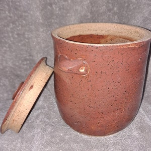 Puede incluir: Un tarro de cer&aacute;mica hecho a mano, de color marr&oacute;n rojizo moteado, con tapa a juego. El tarro tiene forma redondeada, dos asas peque&ntilde;as y un borde de color m&aacute;s claro. La tapa est&aacute; ligeramente entreabierta.