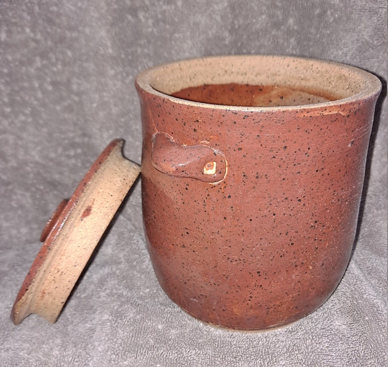 Puede incluir: Un tarro de cer&aacute;mica hecho a mano, de color marr&oacute;n rojizo moteado, con tapa a juego. El tarro tiene forma redondeada, dos asas peque&ntilde;as y un borde de color m&aacute;s claro. La tapa est&aacute; ligeramente entreabierta.