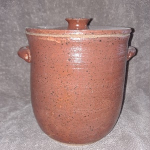 May include: A handmade, speckled, brown ceramic jar with a lid. The jar has two small handles and a rounded base. The lid has a small knob on top for easy removal. The jar is likely used for storage.