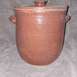 Puede incluir: Una jarra de cer&aacute;mica marr&oacute;n rojizo con tapa, con un patr&oacute;n moteado. La jarra tiene dos asas peque&ntilde;as y un pomo en la tapa. La cer&aacute;mica hecha a mano tiene un aspecto r&uacute;stico y texturizado.