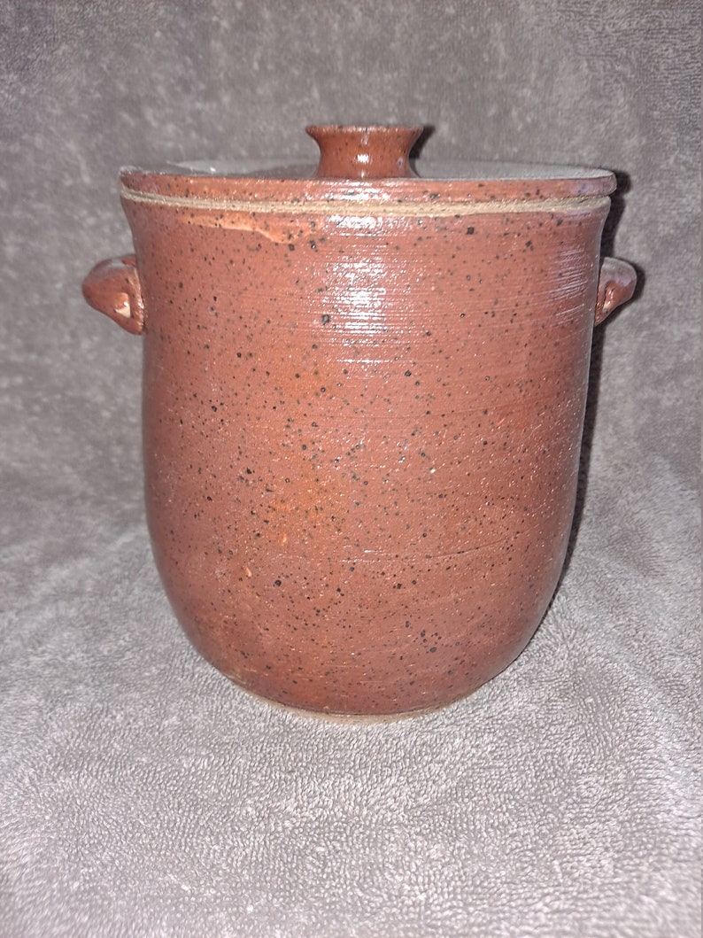 Puede incluir: Una jarra de cer&aacute;mica marr&oacute;n rojizo con tapa, con un patr&oacute;n moteado. La jarra tiene dos asas peque&ntilde;as y un pomo en la tapa. La cer&aacute;mica hecha a mano tiene un aspecto r&uacute;stico y texturizado.
