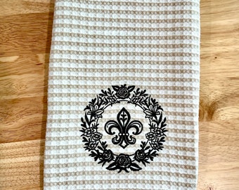 May include: A rectangular waffle-weave kitchen towel featuring alternating horizontal stripes of white and beige. Centered on the lower half is a black embroidered design consisting of a fleur-de-lis symbol surrounded by a circular floral wreath.