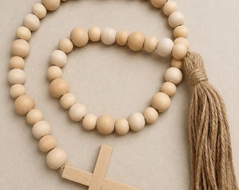 Puede incluir: Una guirnalda decorativa con cuentas de madera redondas, lisas y de color claro ensartadas en un bucle circular. El cordón está adornado con una borla de yute texturizada de color beige y un colgante de cruz de madera clara.