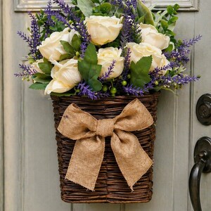 May include: A woven brown basket filled with cream-colored roses, purple lavender, and green foliage, adorned with a large burlap bow. The basket is hanging on a light green door, creating a rustic, welcoming aesthetic.