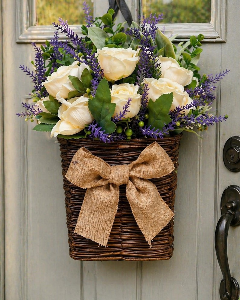 May include: A woven brown basket filled with cream-colored roses, purple lavender, and green foliage, adorned with a large burlap bow. The basket is hanging on a light green door, creating a rustic, welcoming aesthetic.