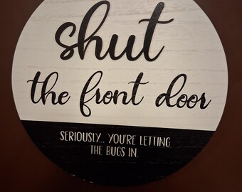 Puede incluir: Un letrero redondo de madera con fondo de veta de madera blanca y una sección inferior negra. El texto dice "shut the front door" en cursiva negra, seguido de "SERIOUSLY... YOU'RE LETTING THE BUGS IN." en letras mayúsculas blancas.