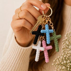 May include: A selection of crochet cross keyrings in black, white, pink, blue, and green. Each cross is attached to a gold-coloured keyring with a clasp. A key is also attached to the ring.