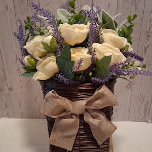 May include: A floral arrangement in a brown woven basket. The arrangement features cream-colored roses, purple lavender, and green foliage. A large burlap bow adorns the front of the basket. The basket is on a small wooden easel.