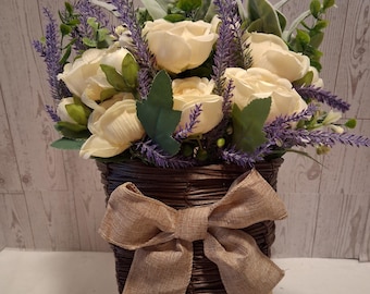 Puede incluir: Un arreglo floral en una cesta de mimbre marrón. El arreglo presenta rosas color crema, lavanda púrpura y follaje verde. Un gran lazo de arpillera adorna la parte delantera de la cesta. La cesta está sobre un pequeño caballete de madera.
