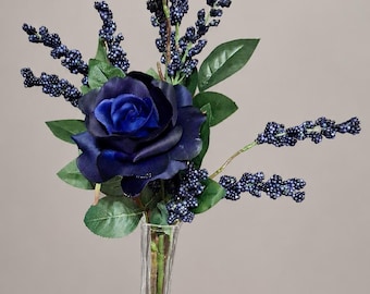 May include: A deep blue rose with green leaves and several stems of small dark blue berries arranged in a clear glass vase. The rose features layered petals in varying shades of navy and royal blue, complemented by textured green foliage.