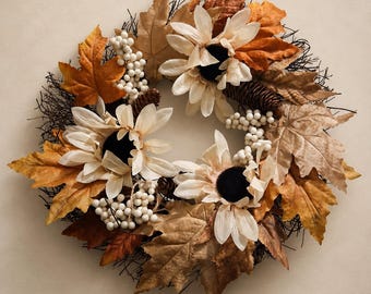 Puede incluir: Una corona circular hecha de ramas de color marrón oscuro, adornada con flores de color crema con centros oscuros, hojas de otoño en varios tonos de naranja y marrón, pequeños racimos de bayas blancas y varias piñas marrones.
