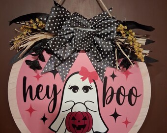 Puede incluir: Un colgador de puerta redondo de madera con fondo rosa y el texto "hey boo" en negro. En el centro hay una ilustración de un fantasma blanco con un lazo rojo y una pequeña calabaza roja. Un lazo negro con lunares blancos corona la pieza.